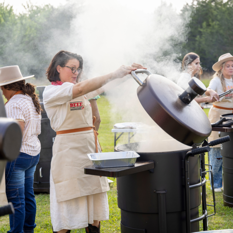 A woman wearing an apron opens a smoker and is engulfed in smoke.